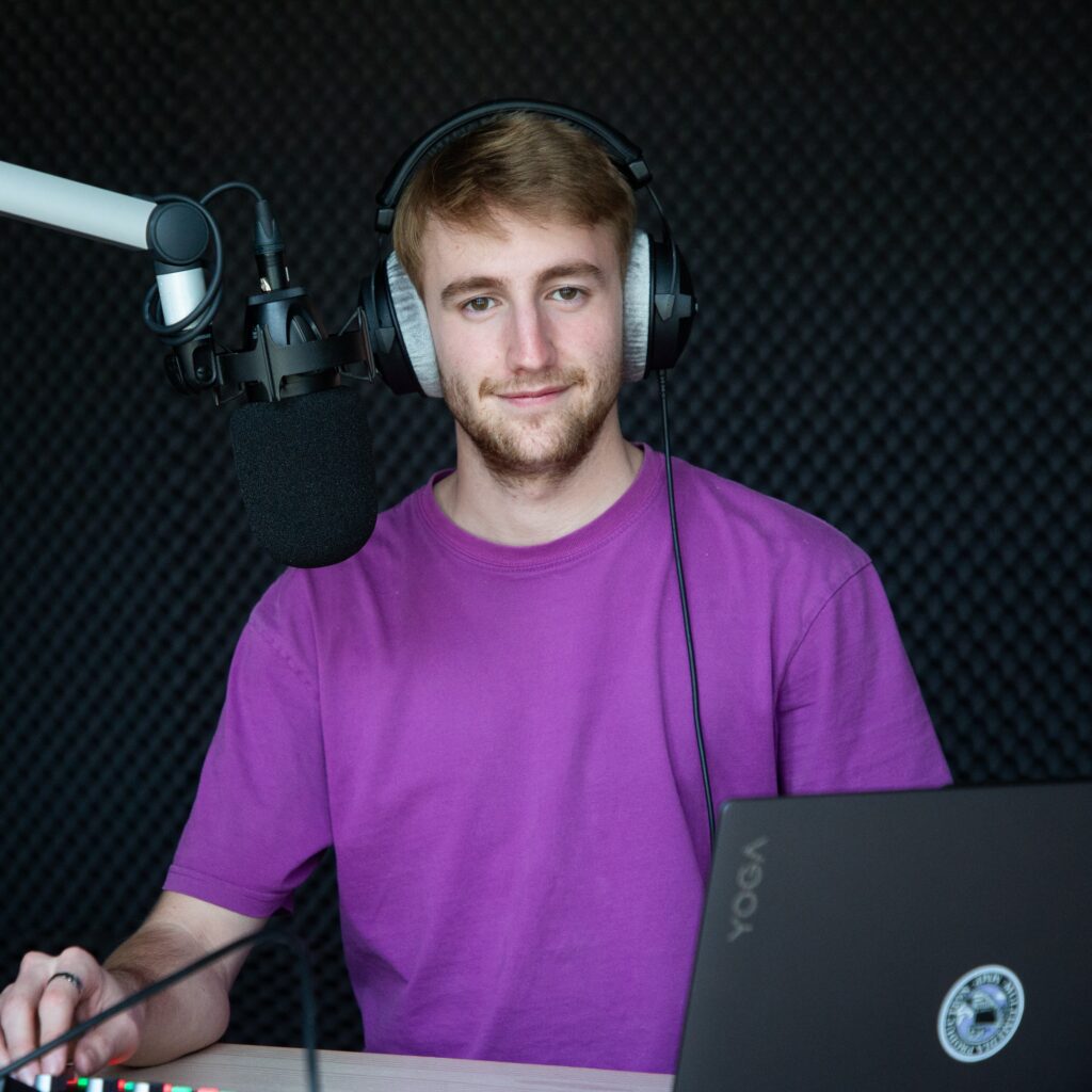 Moritz sitzt mit Kopfhörern auf dem Kopf vor einem Mikrofon in einem Radiostudio. Vor ihm steht sein Laptop und seine Hand bedient das Audiomischpult.